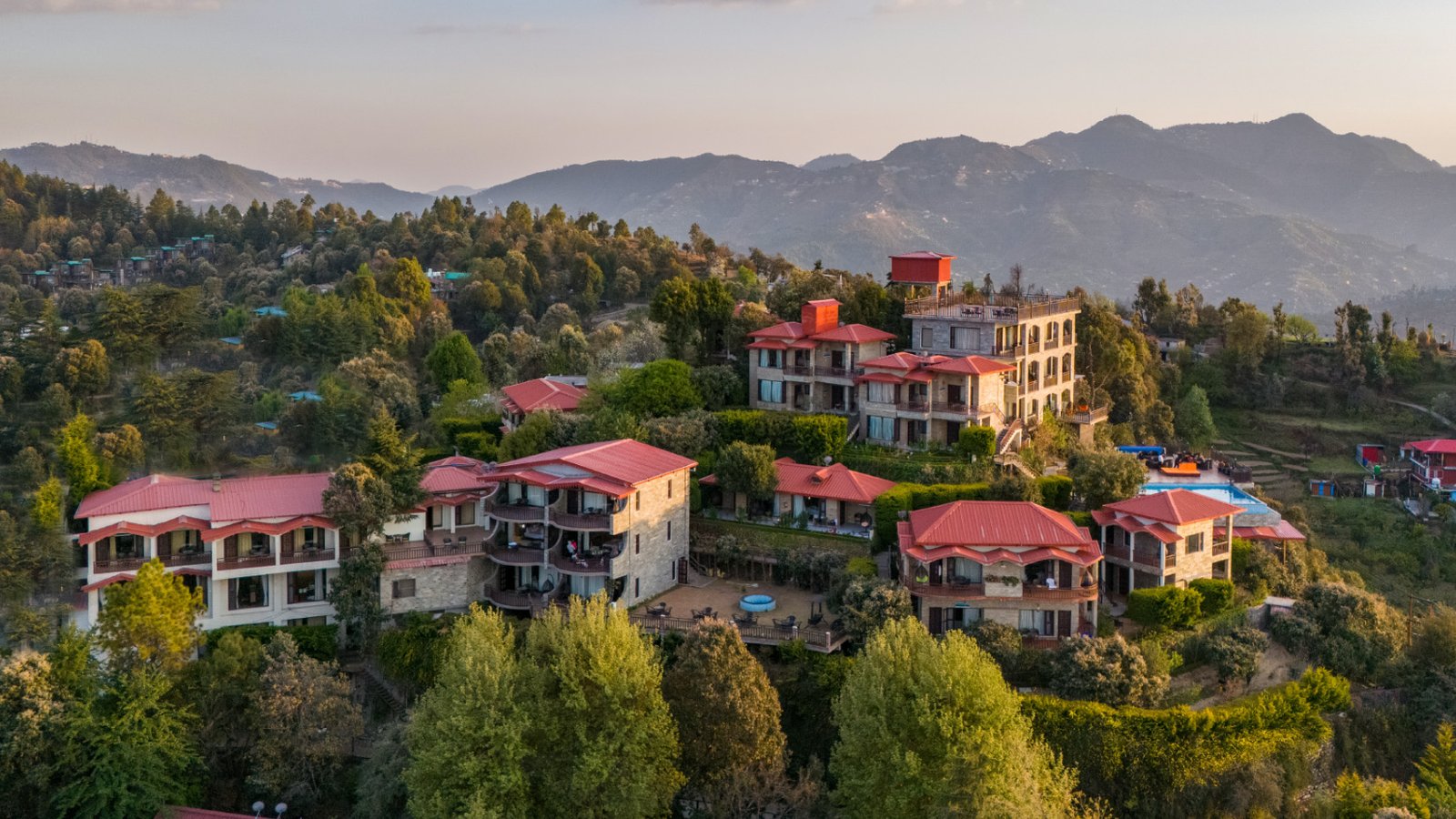 Aerial view of a sprawling luxury resort with several red-roofed stone cottages nestled on a green hillside in the Himalayas.